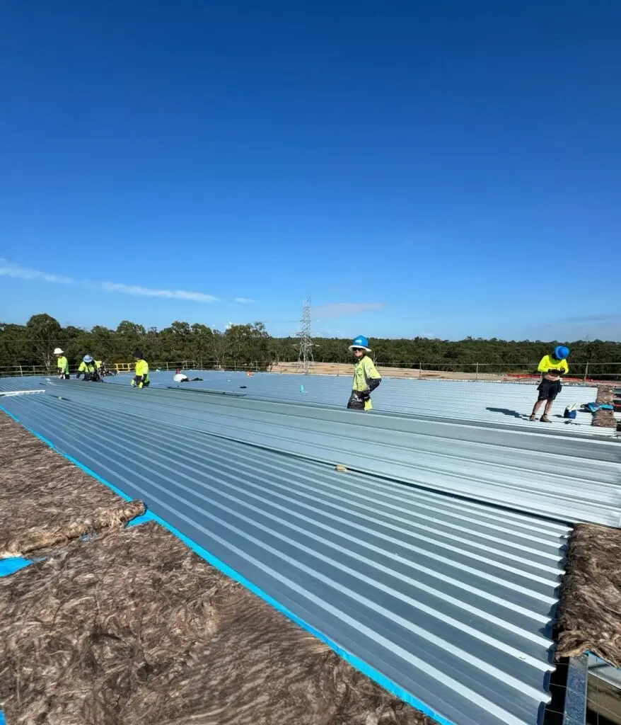 Roofing insulation being installed under corrugated metal sheets on a commercial roof, with workers securing the structure.